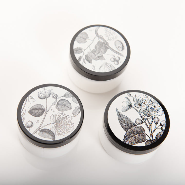 Three white cosmetic jars with black lids featuring botanical illustrations. The jars are arranged in a triangular formation on a plain white surface. Each jar lid has a unique black and white design showing various plant drawings.
