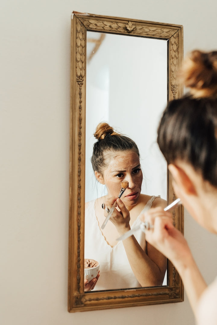 Woman with a bun applying makeup under her eyes using a brush, looking into an ornate framed mirror hanging on the wall. She holds a small bowl in her other hand, reflected in the mirror. The scene is set in a bright, well-lit room.