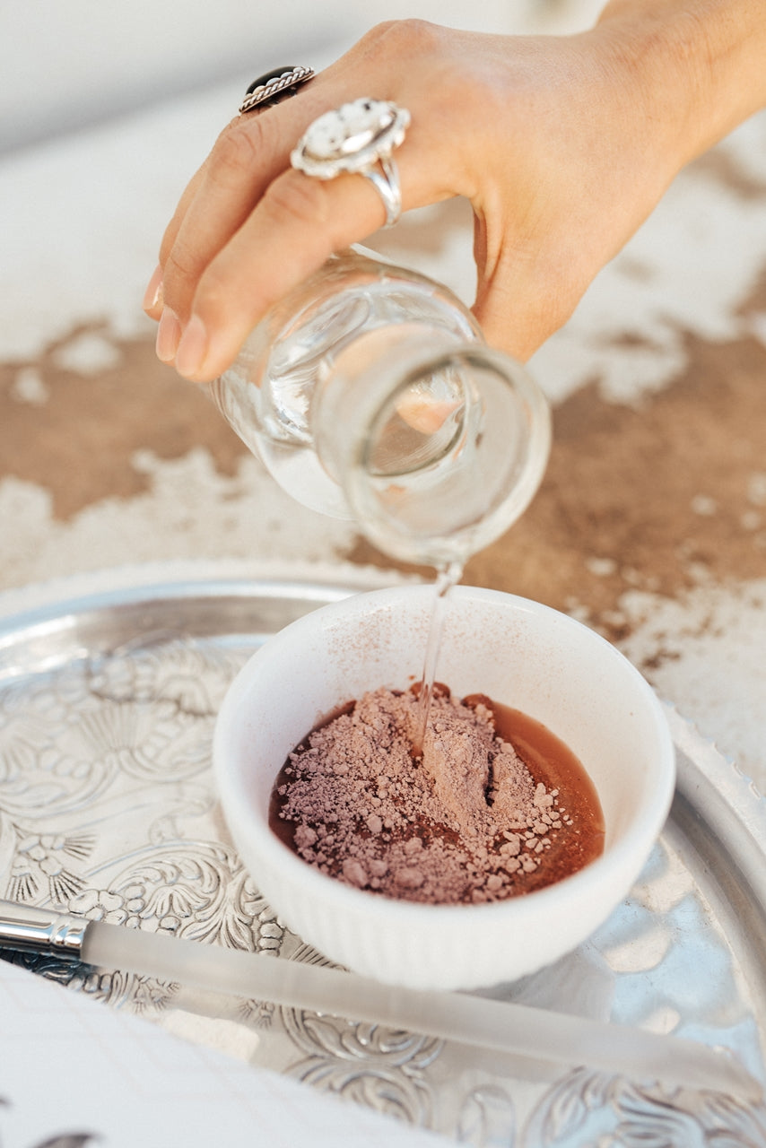 A hand with several rings pours clear liquid from a small glass bottle into a white bowl containing a brown powder. The bowl is placed on an ornate silver tray.
