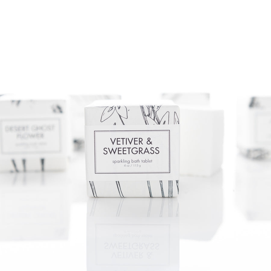 A sparkling bath tablet packaged in a white box with minimalist black botanical illustrations is labeled "Vetiver & Sweetgrass." The background is white, featuring additional similar items slightly blurred.